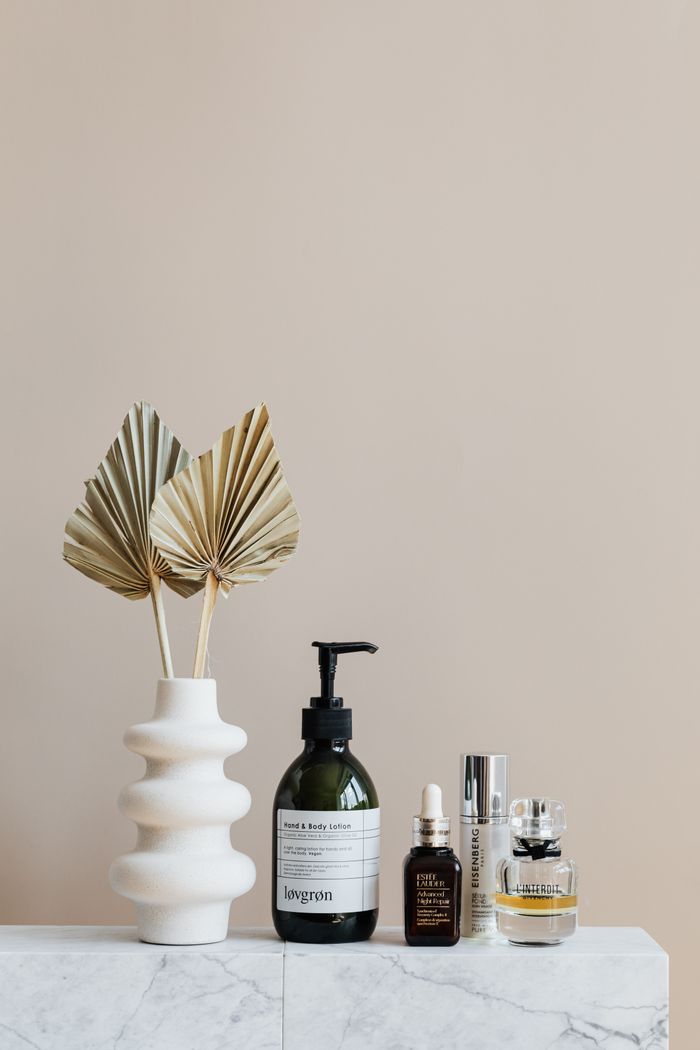 gallery-05 Assorted bottles with cosmetics products placed with creative geometric vase with artificial leaves on white marble shelf against beige wall in modern bathroom interior