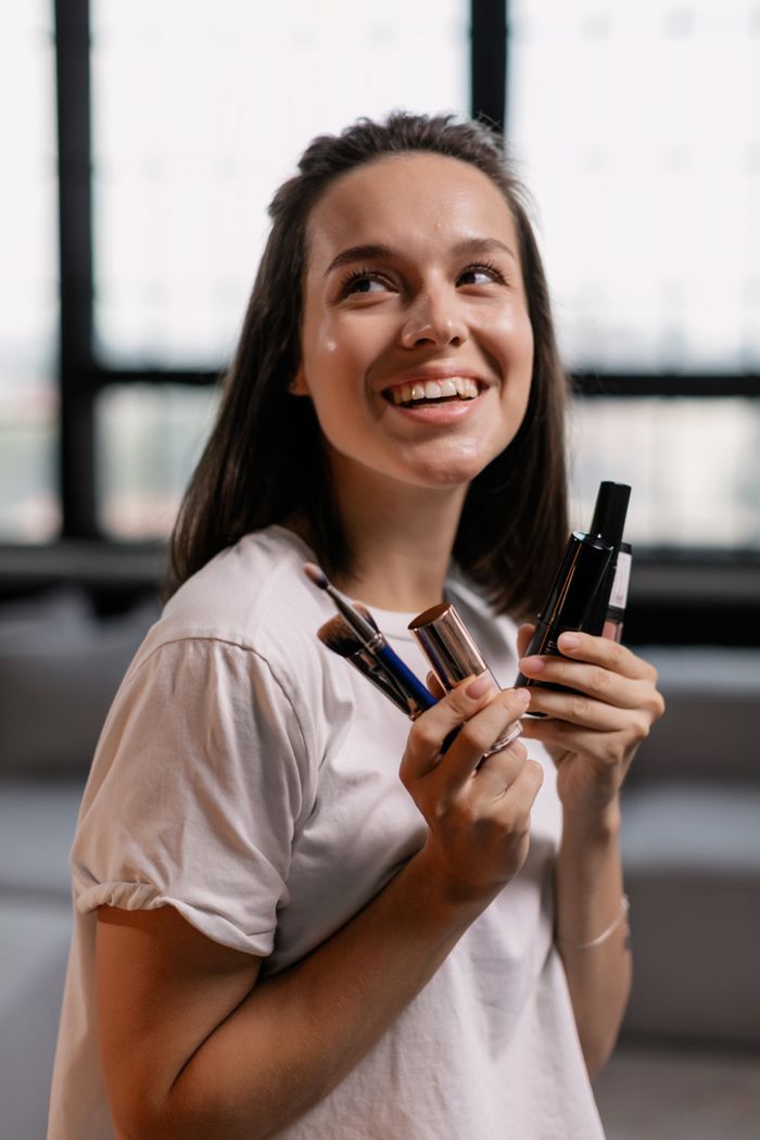 gallery-03 Happy woman indoors holding makeup brushes and products, showcasing beauty routine.